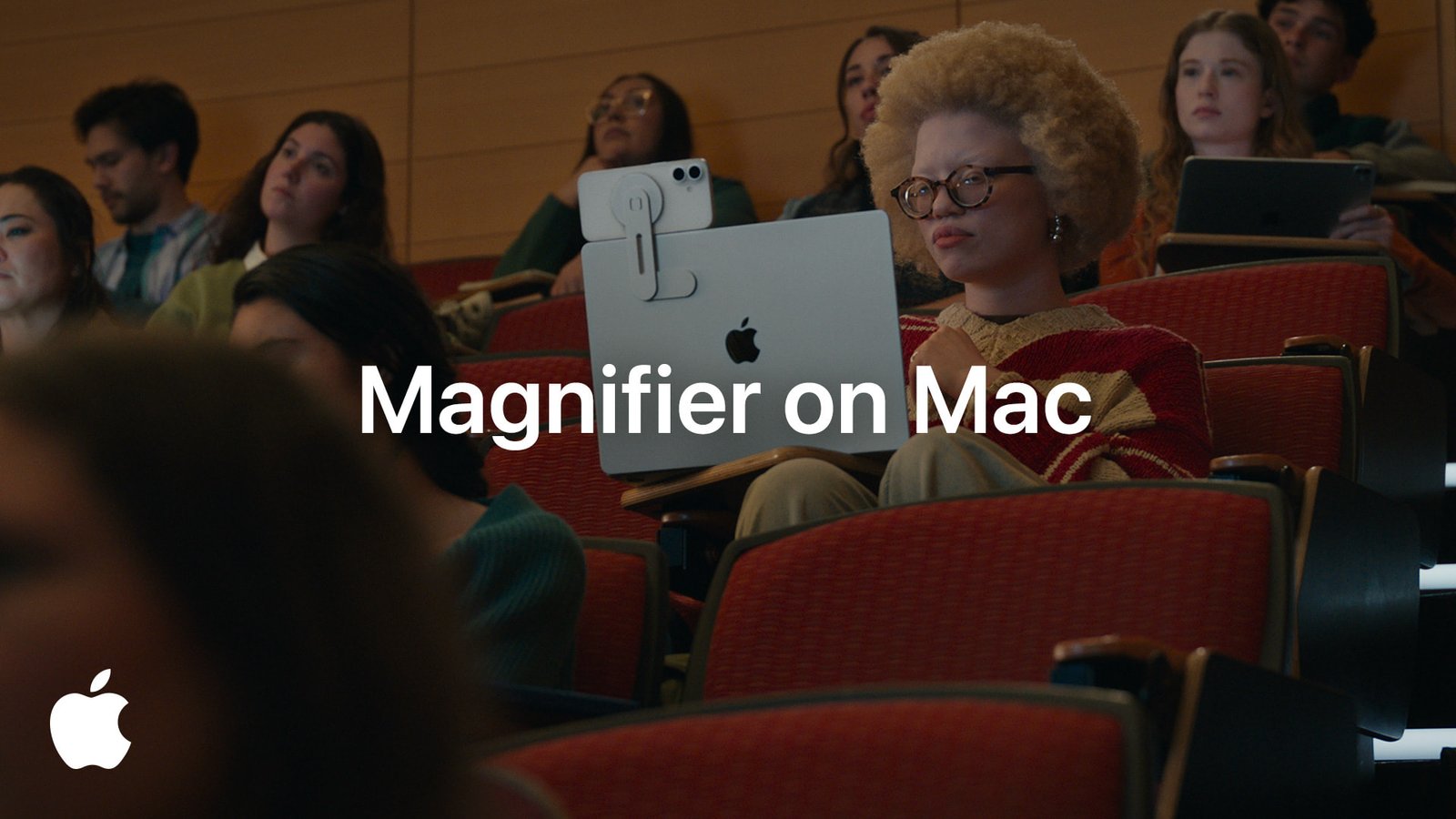 Against the background of red seating, several university students are working and one of them is working on a Mac using the magnifier. She is a female with curly hair and glasses.