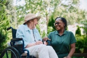 An elderly woman wearing a straw hat, sitting in a wheelchair is being attended to buy healthcare, dressed in teal kneeling at her side
