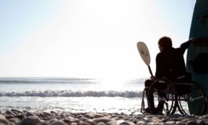 Man is sitting on the beach of the ocean and his wheelchair with a paddle and his hand and his surfboard behind him looking into the sun