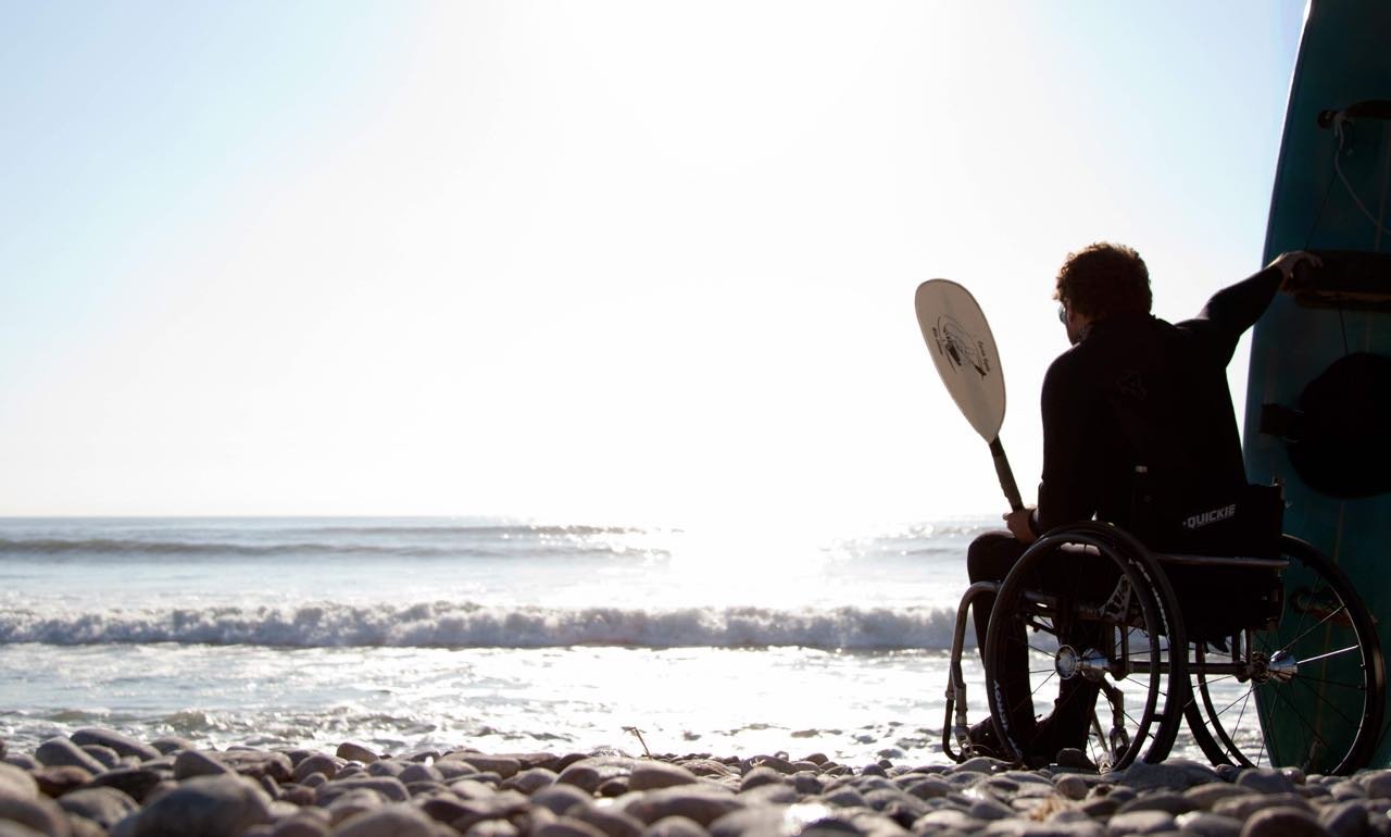 Man is sitting on the beach of the ocean and his wheelchair with a paddle and his hand and his surfboard behind him looking into the sun