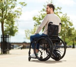 Mad dressed in baby shirt and black pants is sitting in his wheelchair outdoors, beautiful blue skies with some clouds and some trees in the background