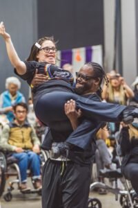 Show as a person with a disability, doing a dance routine with another person in a wheelchair in front of an audience