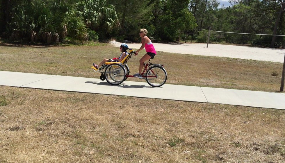 Woman is cycling riding an adaptive bike with a disabled child is sitting in the front of it in a park trail