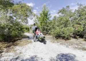 A man is pushing a child and an adoptive three wheel push tricycle on a trail with trees and blue sky in the background