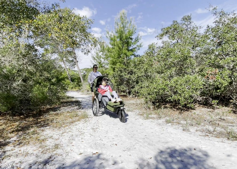 A man is pushing a child and an adoptive three wheel push tricycle on a trail with trees and blue sky in the background