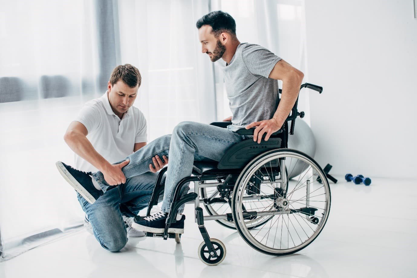 Man, dressed in gray is sitting in a wheelchair with his leg out, and it is being worked on by a physiotherapist male dressed in white