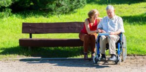 Two persons are out of park bench one is in a wheelchair and they are looking at a map just off the trail with the green grass in the background