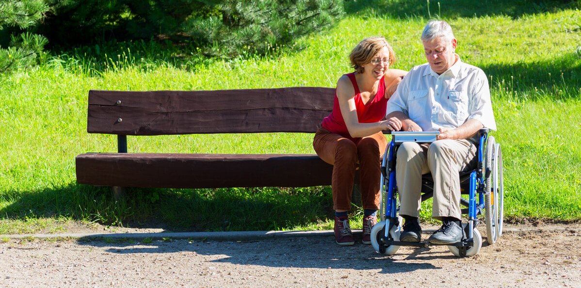 Two persons are out of park bench one is in a wheelchair and they are looking at a map just off the trail with the green grass in the background