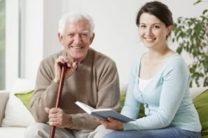Elderly male, patient dressed in brown, sitting on a couch, resting on his cane being intended to buy a female healthcare, dressed in blue