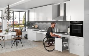 Middle-aged man, dressed in gray in a wheelchair is using the accessible cabinets and his all white kitchen