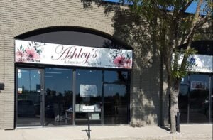 Storefront of building with a sign that says Ashleigh’s apparel and the sidewalk runs across the front of it