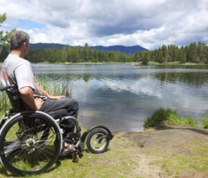 Young man is sitting in his wheelchair with the added front large wheel that enabled him to roll to the Shores edge of a pond with the cloudiest guys in the background