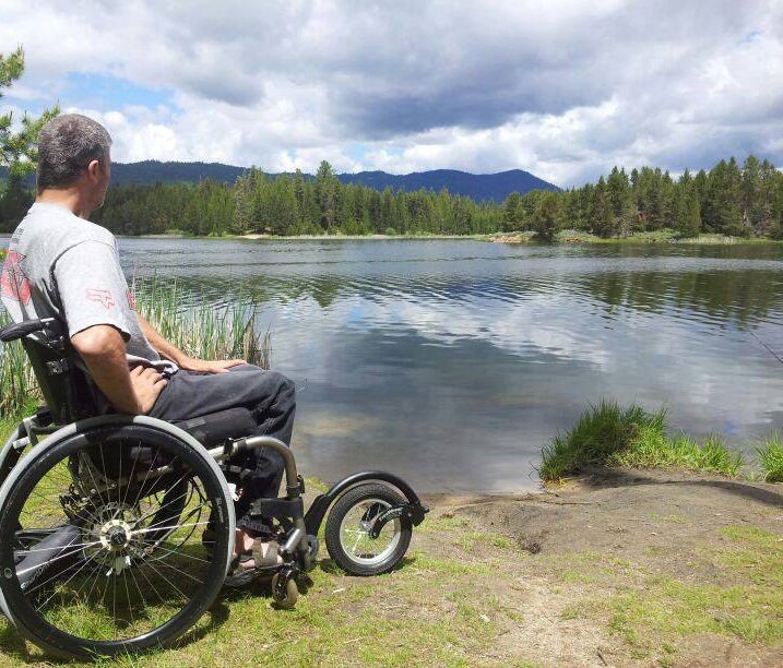 Young man is sitting in his wheelchair with the added front large wheel that enabled him to roll to the Shores edge of a pond with the cloudiest guys in the background