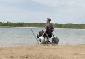 Young man with a disability is sitting in a beach wheelchair on the edge of the sand and water at a lake