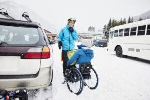 Women dressed in blue is sitting in a wheelchair in the snow on a street trying to get through one side. There’s a white bus the other side there’s a car parked. Someone is trying to help the woman.