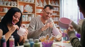 A man and a woman are painting pottery projects on a table, filled with other colourful clay projects and brushes and paints with a shelf behind them full of more pottery projects