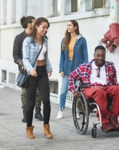 A group of four young adults walking on a city sidewalk. One woman in a denim jacket and another in a gray top and black jeans are smiling as they walk towards a man in a wheelchair wearing a plaid jacket. The setting is urban.