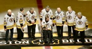 A group of seven men, including a Boston Bruin in a wheelchair, wearing yellow and black jerseys, gathered on an ice rink for a ceremonial event. They are standing on a logo representing the Boston Bruins.