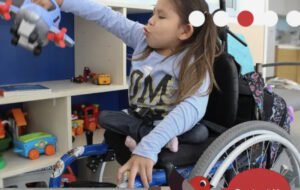 A young girl in a wheelchair playing with a toy airplane in a brightly lit room filled with colorful toys, showcasing interactive play at a Rehab Centre Community Recreation.