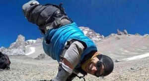 A man with no legs is hanging onto an airplane, wearing a blue vest and goggles with clouds and blue sky in the background