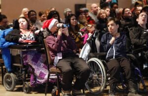 A diverse group of people, including individuals in wheelchairs, seated in an audience at an event. One person wears a Santa hat and holds a camera, while others are engaged in the presentation. The setting suggests a communal gathering, promoting inclusivity.