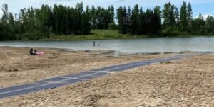 A sandy beach area with a blue accessibility mat leading to a calm body of water. In the background, people are enjoying the water, and trees line the shore, providing a natural setting. The image highlights accessibility mats on Manitoba beaches.