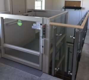 A CHCP elevator lift installation in a modern interior, featuring a glass enclosure and a wooden handrail. The elevator is positioned against a wall with sunlight coming through nearby windows, providing a clear view of the lift's design.