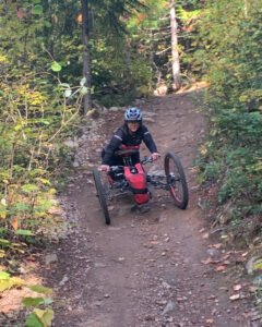 A person using a handcycle on a dirt trail surrounded by trees. The individual is wearing a helmet and is positioned on the handcycle, navigating through a forested path. The image highlights adaptive cycling, relevant to National Trail Guidelines - Trans Canada Trails.