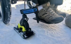Close-up view of a wheelchair equipped with a yellow snow sled attachment on a snowy surface. The user's shoes are visible, showing traction tread suitable for outdoor conditions. This image relates to Accessible Sport - Events List.