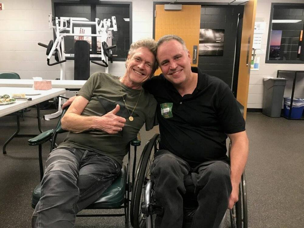One man sitting in his wheelchair and another man sitting beside him, who’s lost his arm, and they are both professional drummers sitting in a room