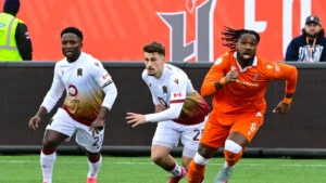 to valour football club players in white sweaters, and white shorts are chasing after opposing player wearing bright orange and long hair and the ball