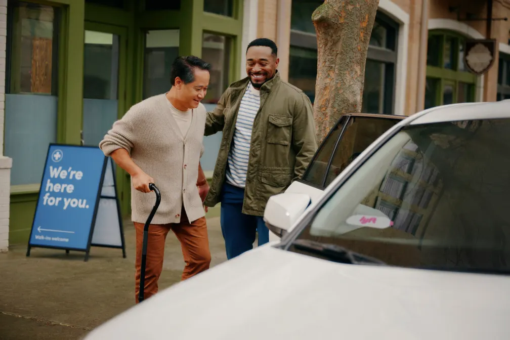LYFT driver is holding the door open up his white car well an elderly gentleman wearing a sweater and using a cane is going towards the open door