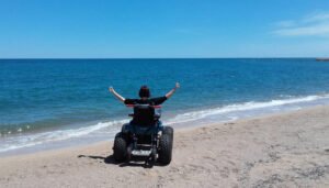 Young man in a wheelchair with beach tires is waving arms in the air, looking at the large expanse of blue ocean while he’s on the water edge on the sand