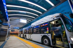 A large welder bus garage has a double length, transit bus, white, and blue FaceTime Adam angle towards the front