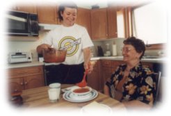 Woman seated at the kitchen table is being assisted by a healthcare aid standing wearing white
